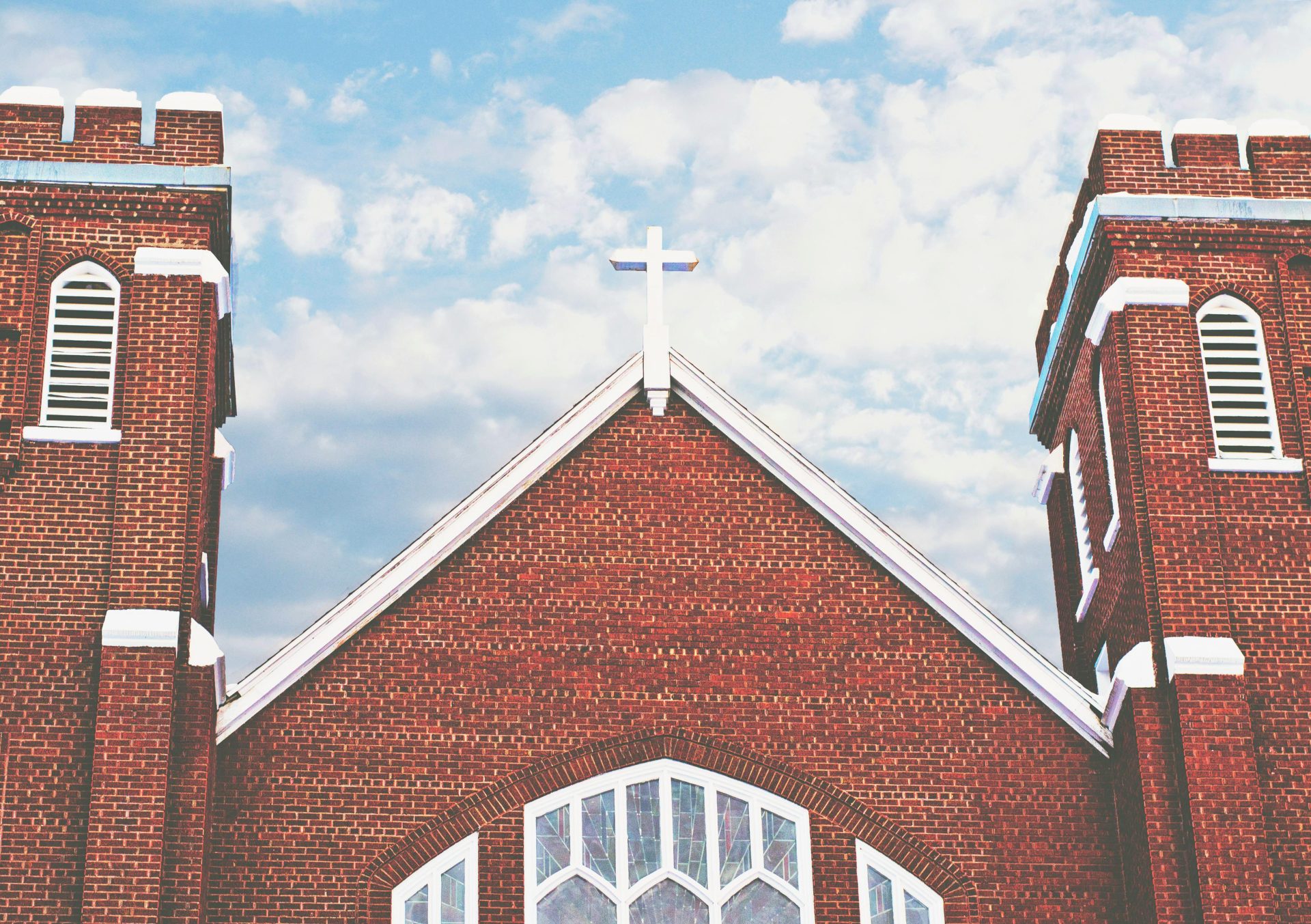 Red brick church with a cross and arched windows under a blue sky with clouds.